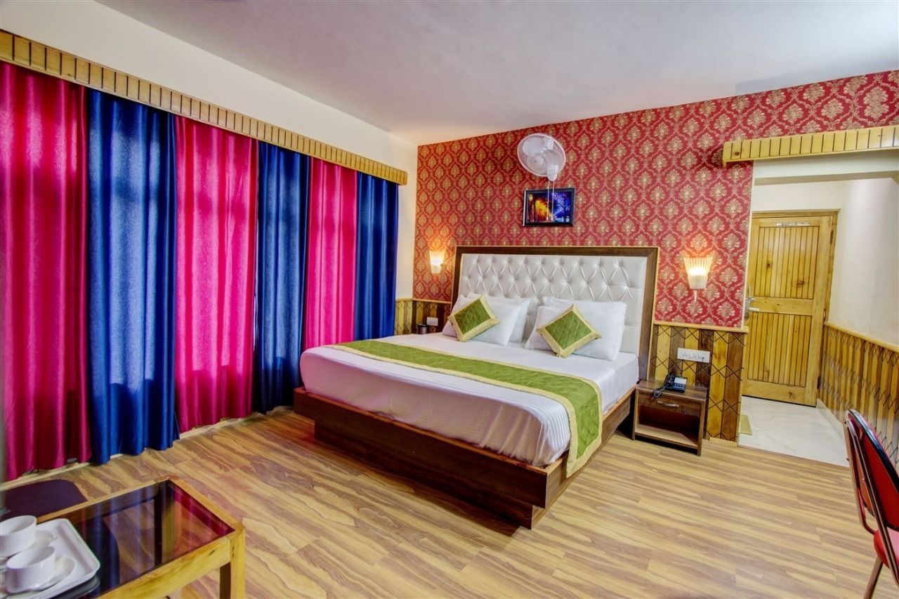 Ein Hotelzimmer mit einem großen Bett, Holzboden, Vorhängen, einem Nachttisch, einem Sessel und einem Wandventilator.