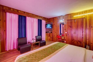Quarto de hotel com lambris de madeira, uma cama, duas cadeiras, uma mesa pequena, uma televisão e uma cadeira vermelha.