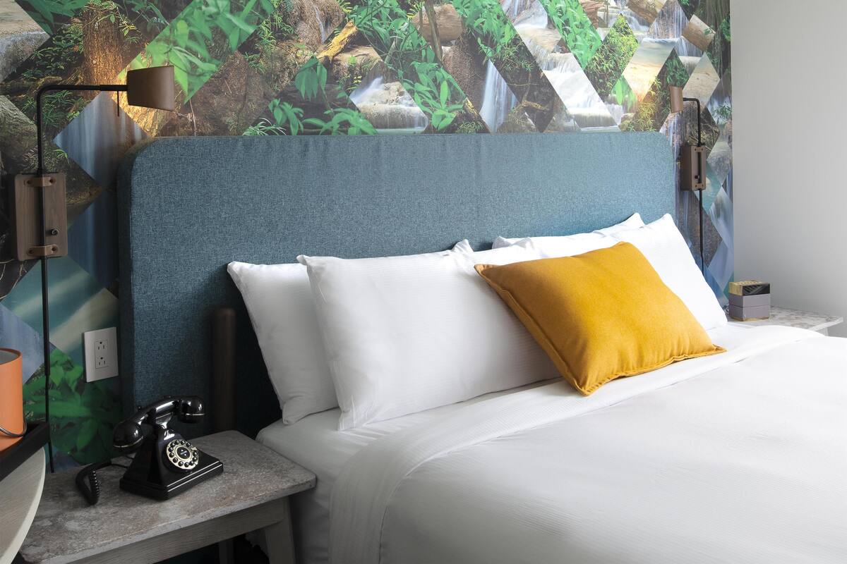 A bed with white and mustard pillows, a nightstand with a rotary phone, and a wall with a nature-themed mural.