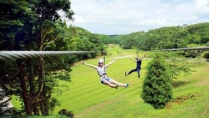Two people ziplining over a lush green landscape with trees and a clear sky. - Hotel Swan Lake Nagara (Ichihara)