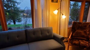 Un salon avec un canapé de couleur sombre, un fauteuil en bois et une lampe sur pied. Il y a une fenêtre avec des rideaux et un tableau au mur.