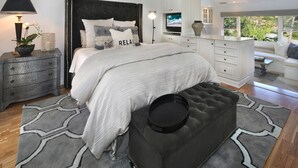 A bedroom with a large bed, a nightstand with a lamp, a TV, and a view of the outdoors. - Serenity in the Hills - A Chic Coastal Apartment with Balcony & Ocean Glimpses (Laguna Beach)