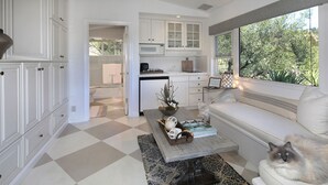 A modern kitchen with white cabinetry, a dining area with a white sofa, and a view of greenery through a large window. - Serenity in the Hills - A Chic Coastal Apartment with Balcony & Ocean Glimpses (Laguna Beach)