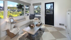 A cozy room with a large window, a wooden bench, a small table with decorative items, and a checkered floor. - Serenity in the Hills - A Chic Coastal Apartment with Balcony & Ocean Glimpses (Laguna Beach)