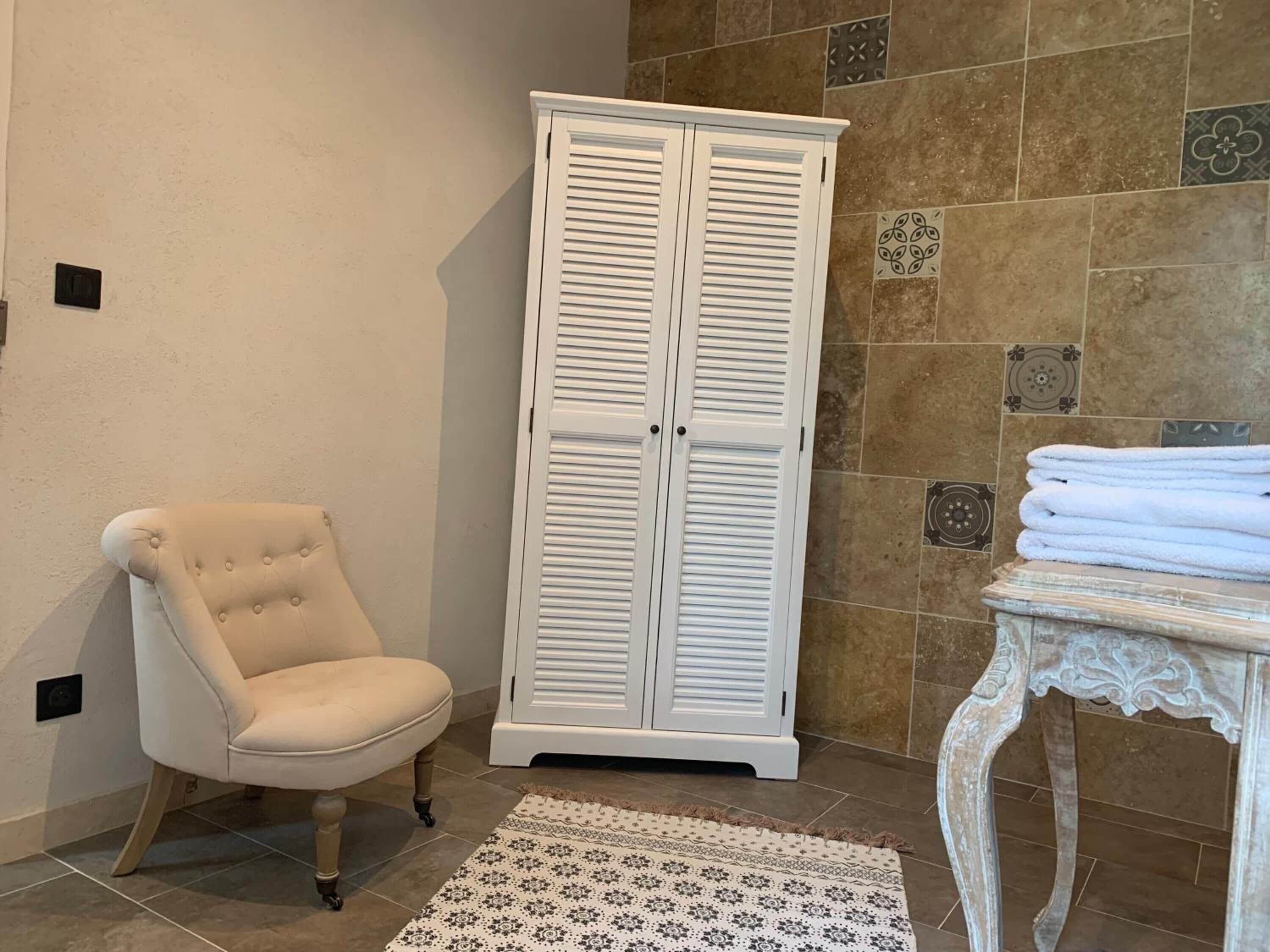 A bathroom with a white vanity, a beige armchair, a white cabinet, and a patterned rug.
