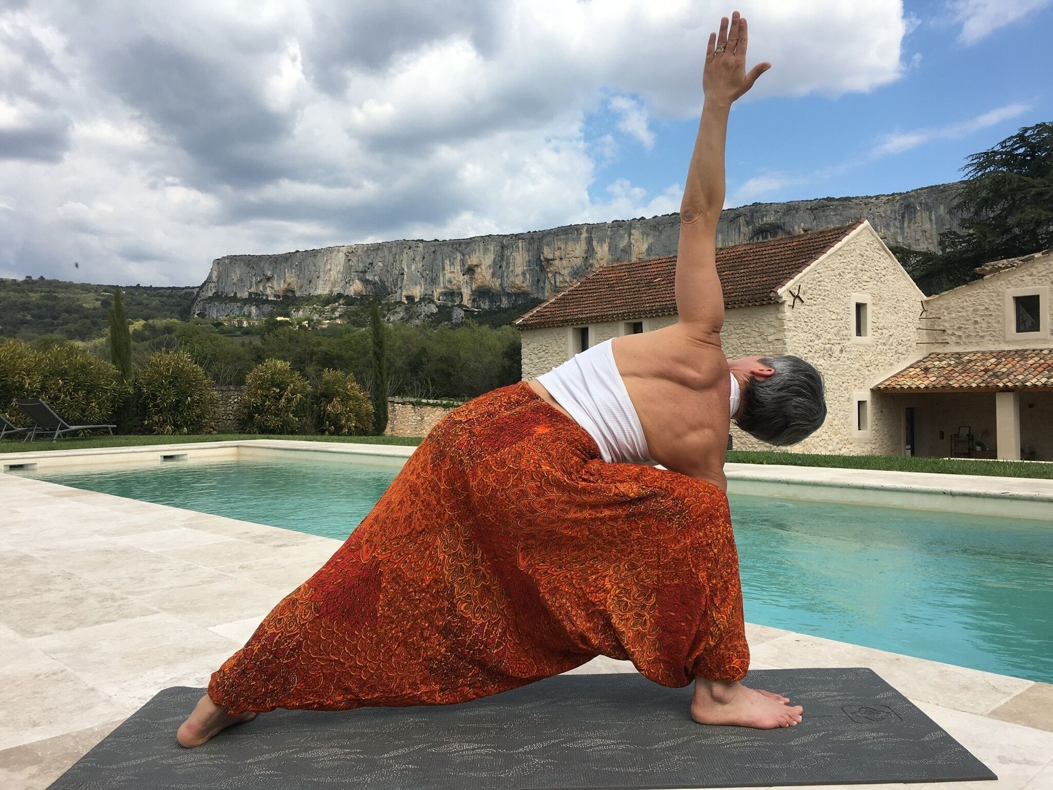 Eine Person führt eine Yoga-Pose auf einer Matte aus, neben einem Pool mit einem Steinhaus und Klippen im Hintergrund.