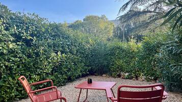 A garden patio with two red metal chairs and a small table, surrounded by dense green foliage and a clear blue sky.