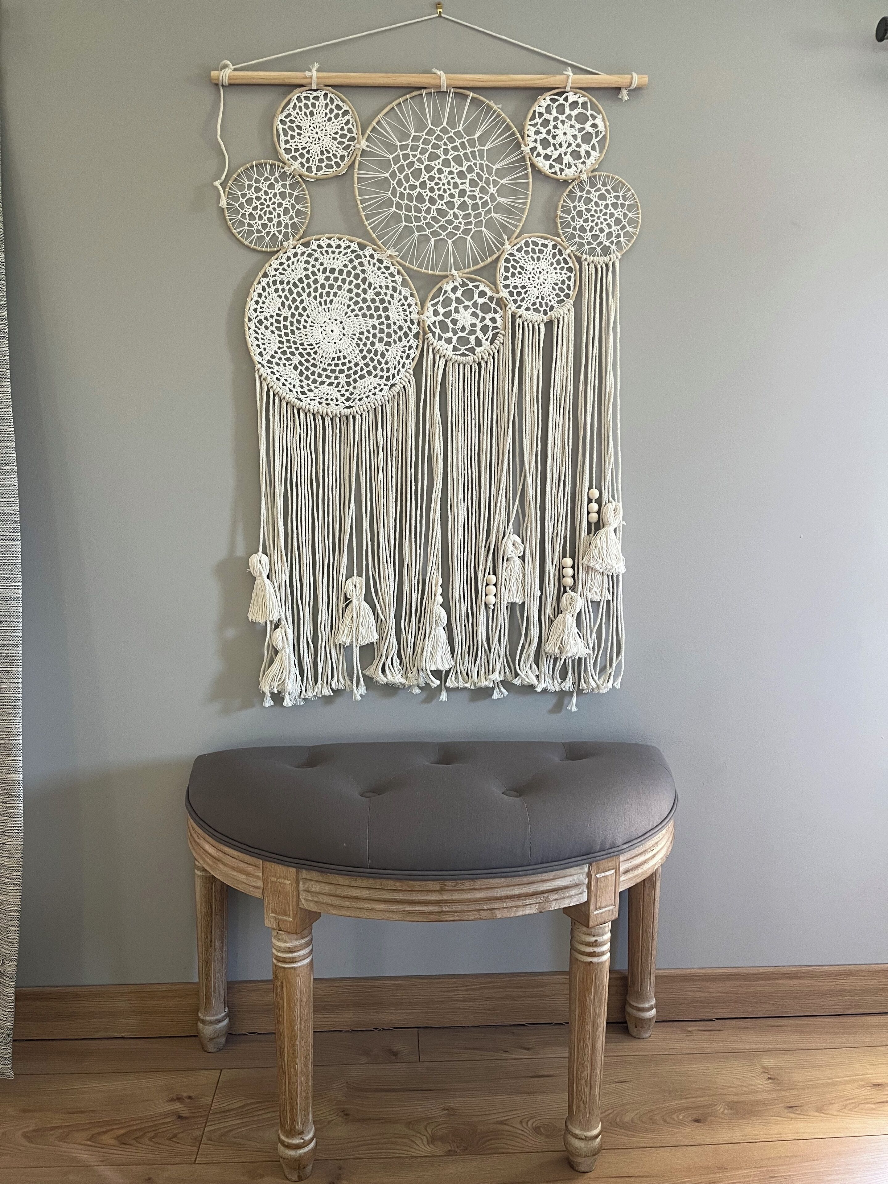 A macrame wall hanging with circular and circular designs, a grey cushioned stool, and wooden floorboards.