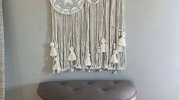 A macrame wall hanging with circular and circular designs, a grey cushioned stool, and wooden floorboards.