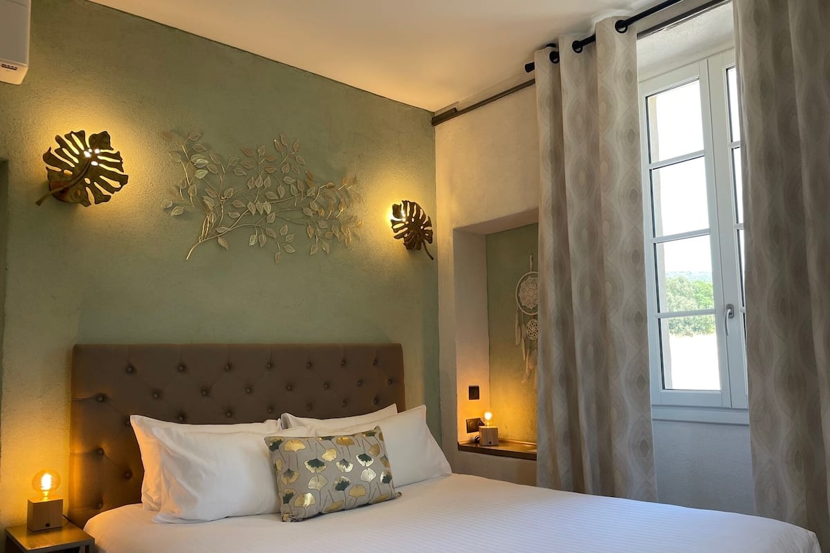 A bedroom with a large bed, a nightstand with a lamp, decorative wall art, and a window with curtains.