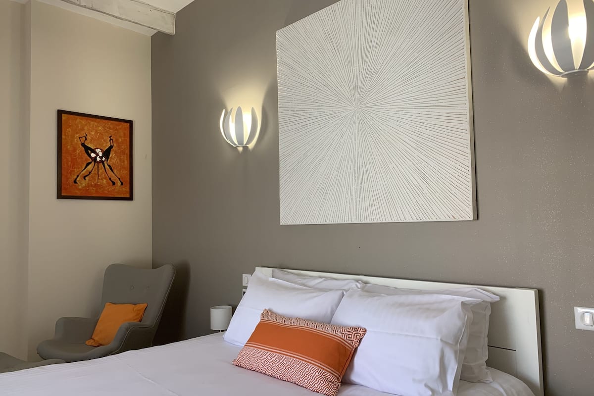 A hotel room with a bed, a chair, a wall-mounted artwork, and two wall sconces.