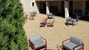 A stone building with a tiled roof, outdoor seating area with chairs and tables, and a gravel ground. - Le Mas aux cedres (Lioux)
