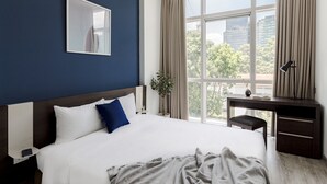 Une chambre d’hôtel avec un grand lit, un bureau, une fenêtre avec des rideaux et une vue sur les bâtiments et les arbres à l’extérieur.