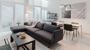 Un salon moderne avec un canapé d’angle gris, une table basse décorée d’oranges et un coin repas comprenant une table et des chaises blanches.