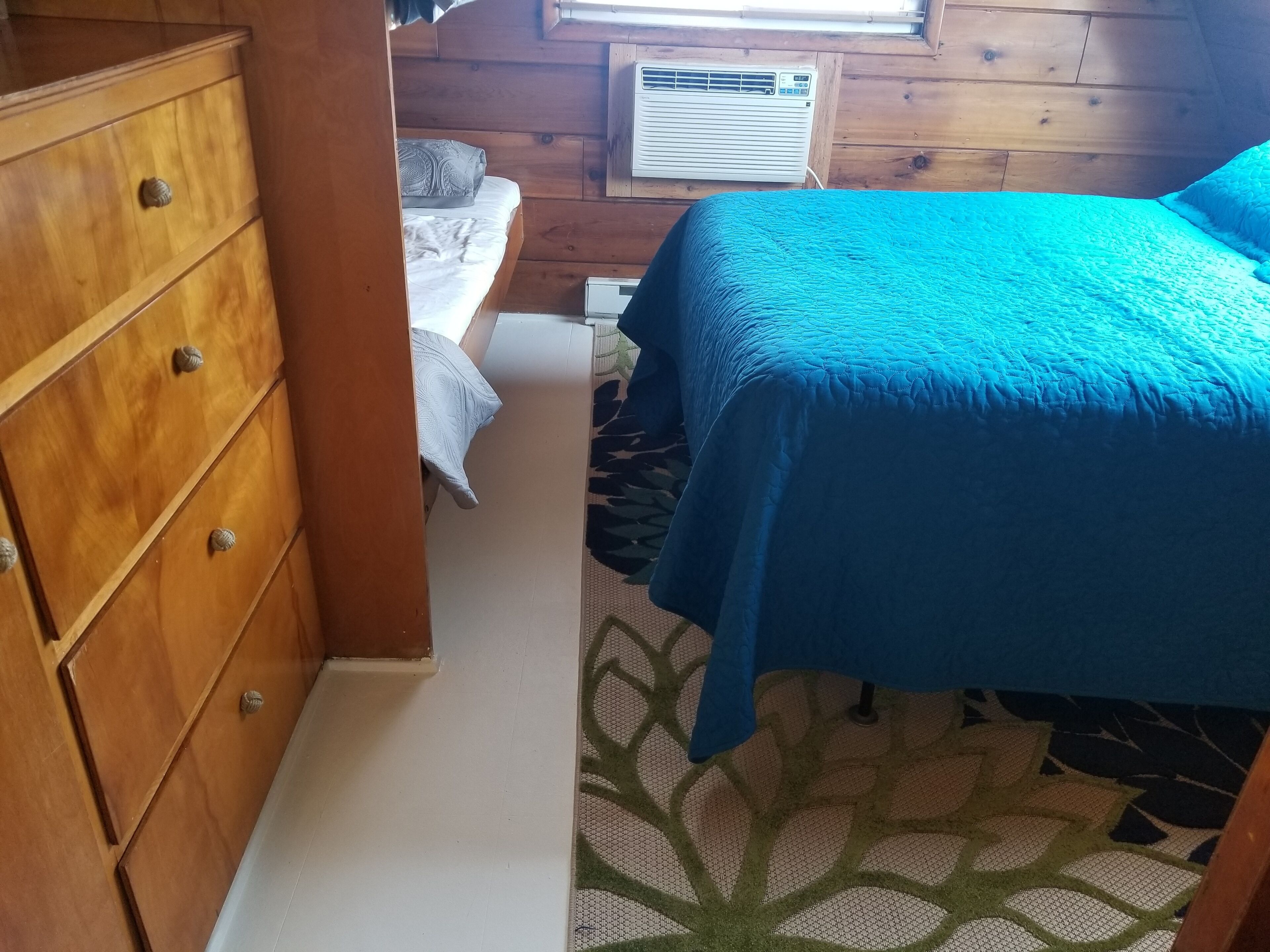 A bedroom with a wooden dresser, a bed with a blue bedspread, a window with blinds, and a wall-mounted air conditioner.