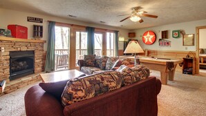 A cozy living room with a stone fireplace, a pool table, and a large window with curtains.