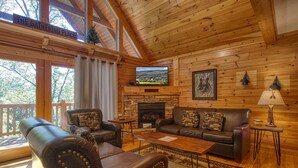 A cozy cabin interior with a stone fireplace, leather sofas, a flat-screen TV, and a wooden deck visible through large windows.