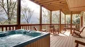 A wooden deck with a hot tub, wooden chairs, and a view of trees and mountains.