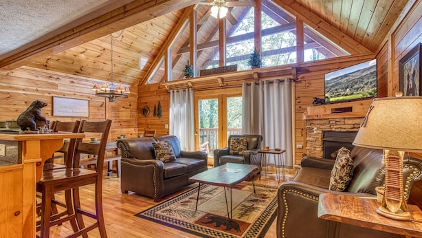 A cozy log cabin interior with a fireplace, leather sofas, wooden floors, and a large window overlooking a scenic landscape.
