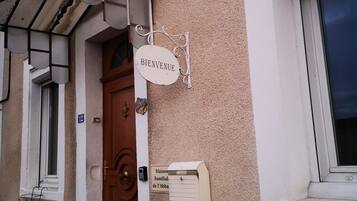 A building entrance with a wooden door, a 'BIENVENUE' sign, a mailbox, and a window with a white frame.