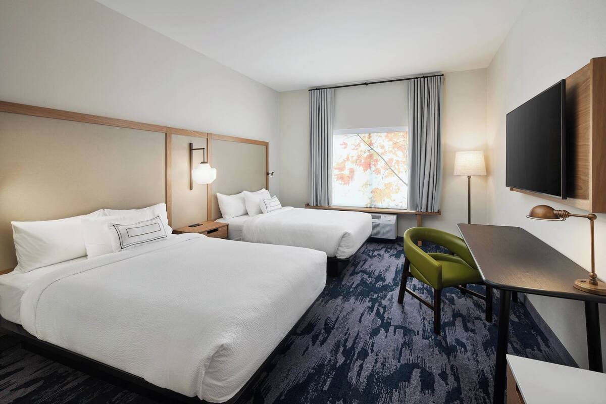 A hotel room with two beds, a desk, a chair, a TV, and a window with curtains.