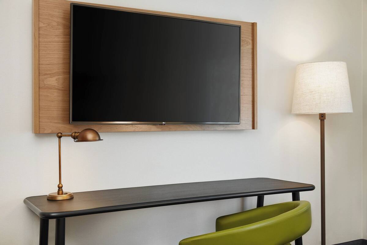 A room with a flat-screen TV mounted on a wooden panel, a desk with a lamp, and a green chair.