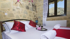 Uma cama com roupa de cama branca e vermelha, um tabuleiro com copos de vinho e uma janela com vista para uma colina.