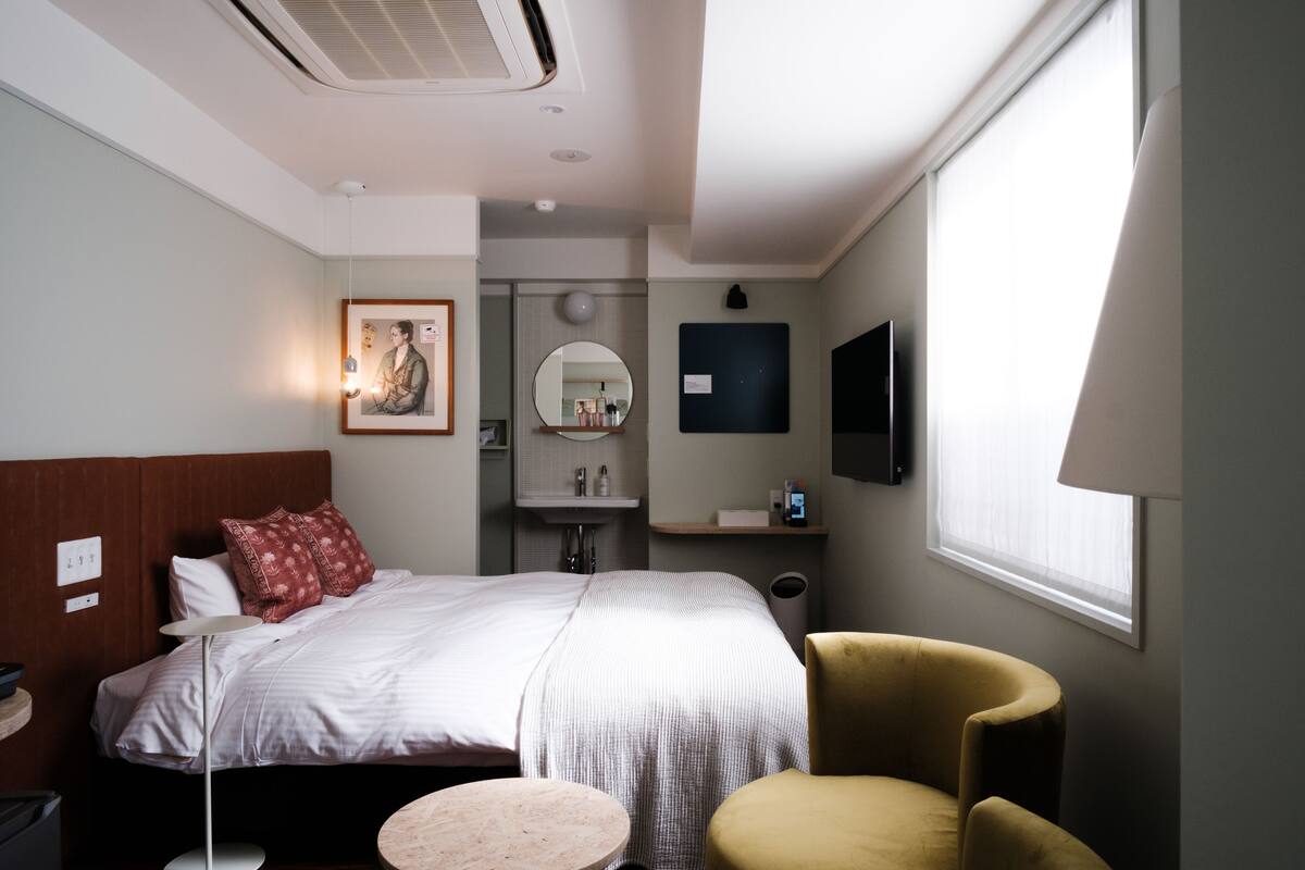 Een hotelkamer met een bed, nachtkastje, stoel, ronde tafel en een aan de muur bevestigde televisie.