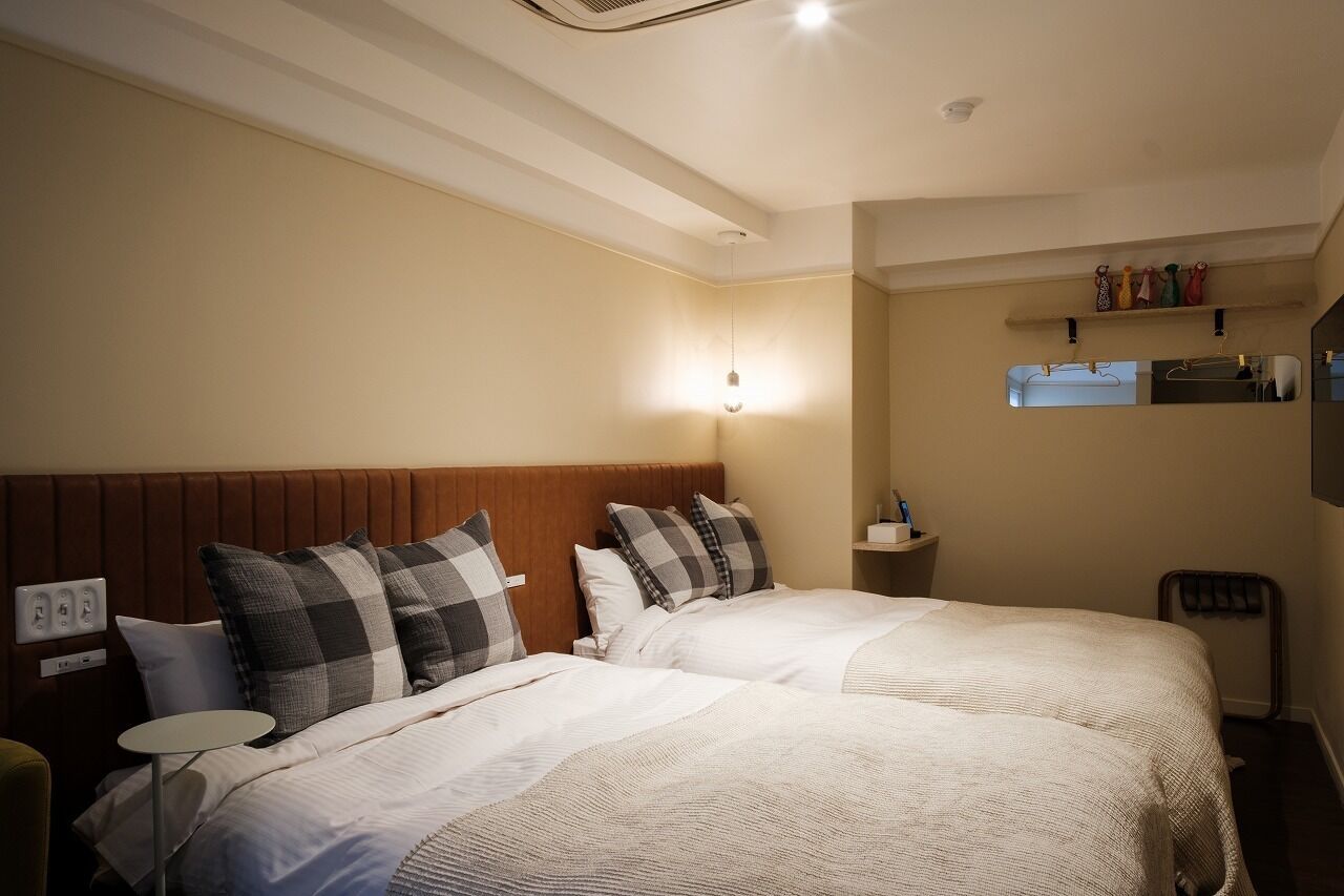 A hotel room with two beds, a bedside table, a wall-mounted shelf with toiletries, and a ceiling light.