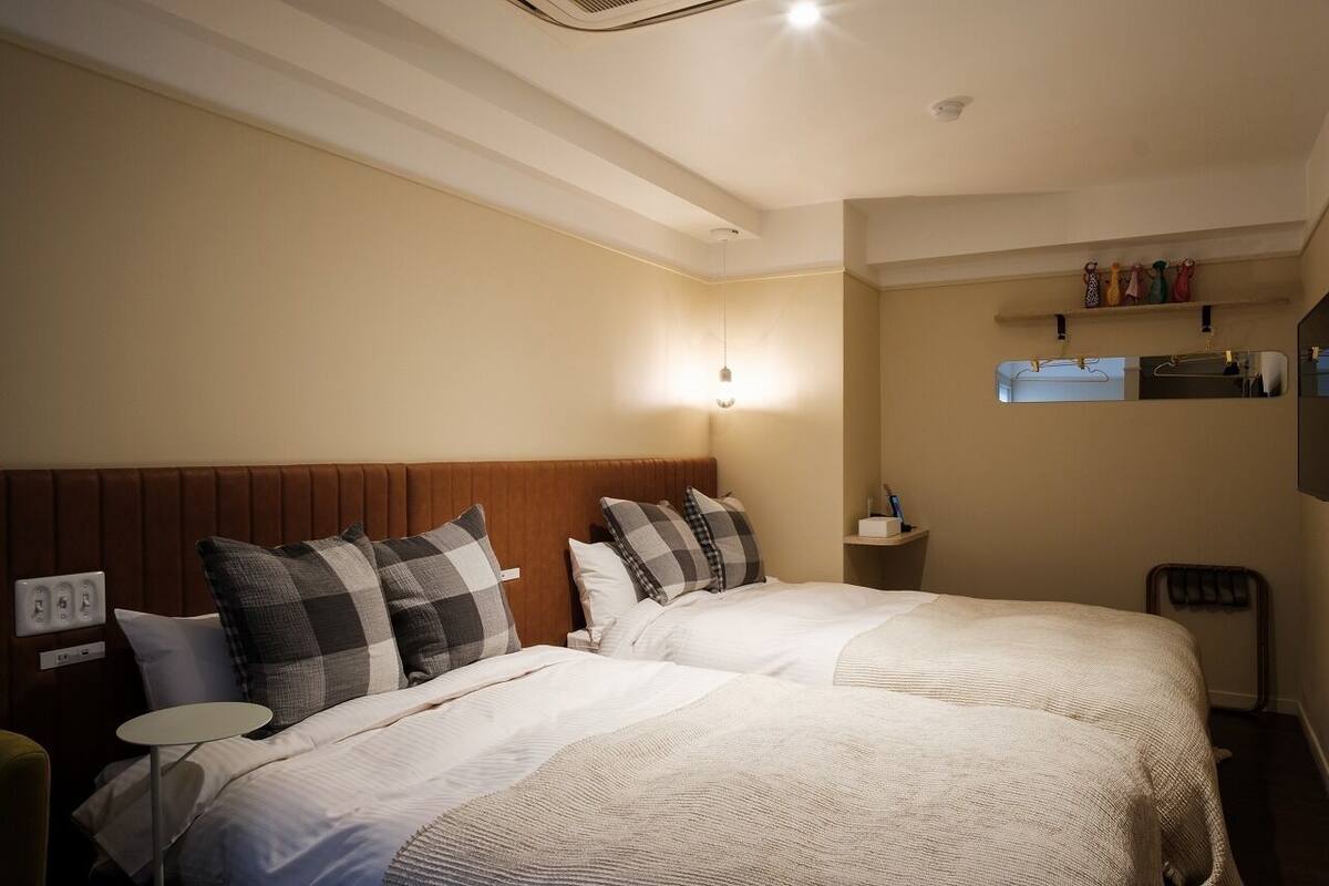 A hotel room with two beds, a bedside table, a wall-mounted shelf with toiletries, and a ceiling light.