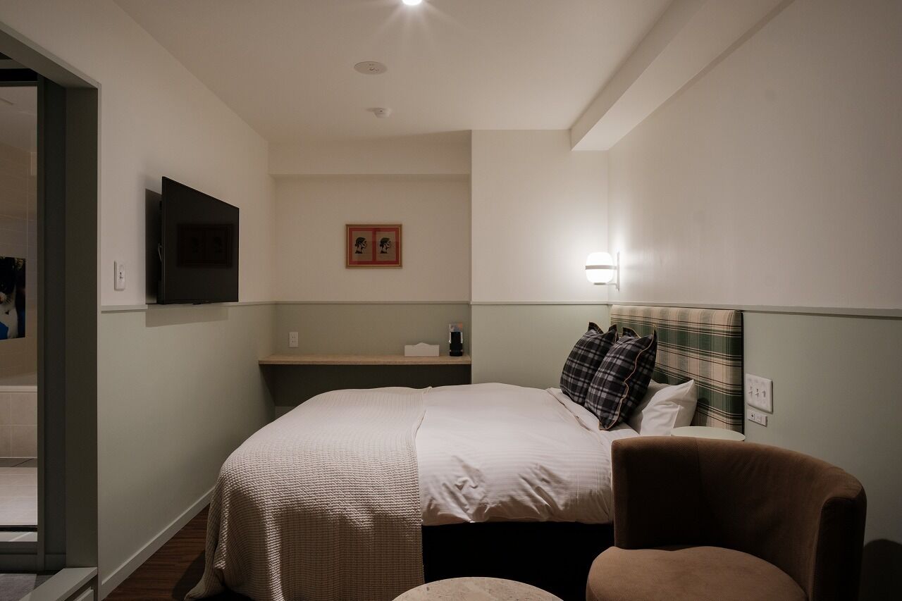 Habitación de hotel con cama, una silla, televisor, mesita de noche y lámpara de pared.