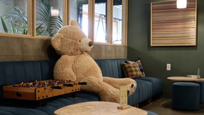 Ein gemütliches Zimmer mit einem großen, beige farbenen Teddybären, der auf einer blauen Couch sitzt, einem Kicker und einem kleinen Tisch mit einem Schild.