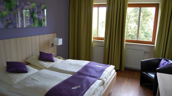 A hotel room with two beds, a desk, a chair, a window with curtains, and a radiator. - Wildkräuterhotel (Windelsbach)