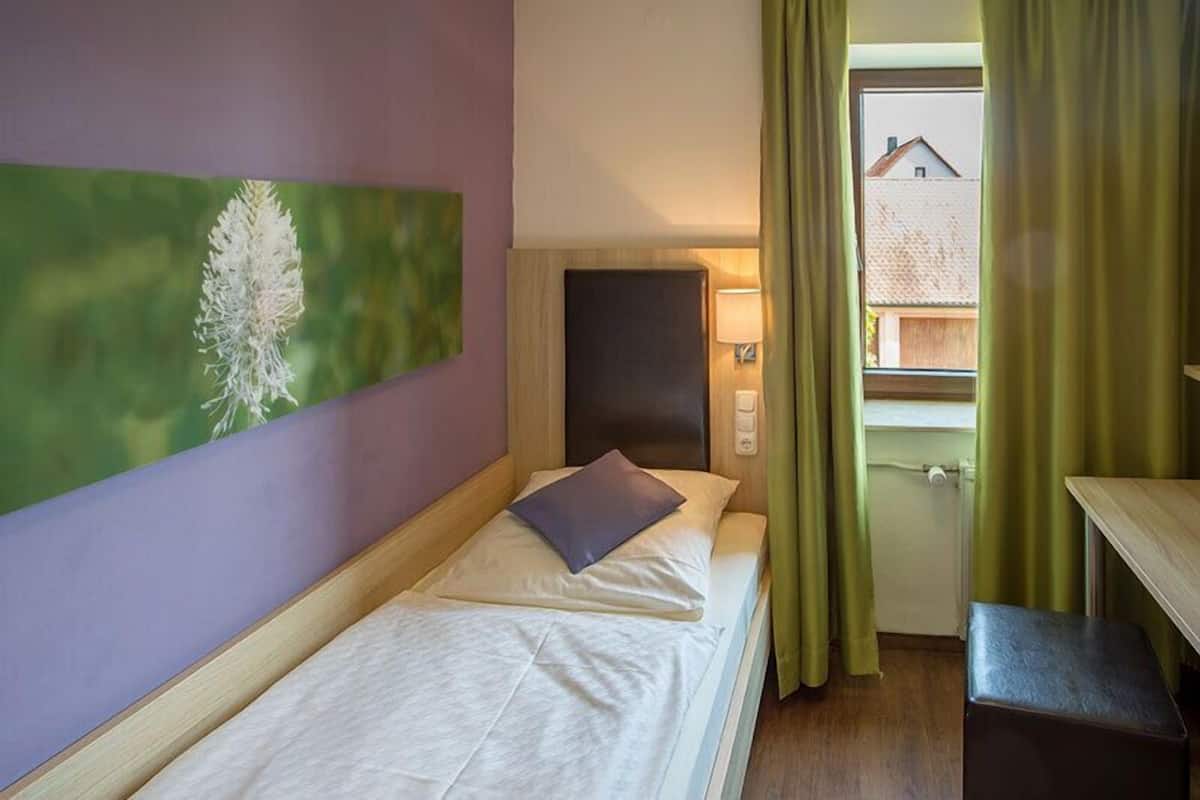 A single bed with a pillow and a headboard, a bedside table, a lamp, and a window with curtains.