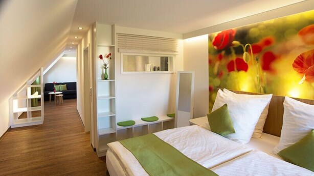 A modern hotel room with a large bed, a green throw, a poppy-themed wall, and a view of the adjacent room. - Wildkräuterhotel (Windelsbach)