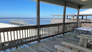 Terraza de madera con barandilla que da a un muelle y al océano. Hay mesas de picnic y un área cubierta con una mesa y sillas.