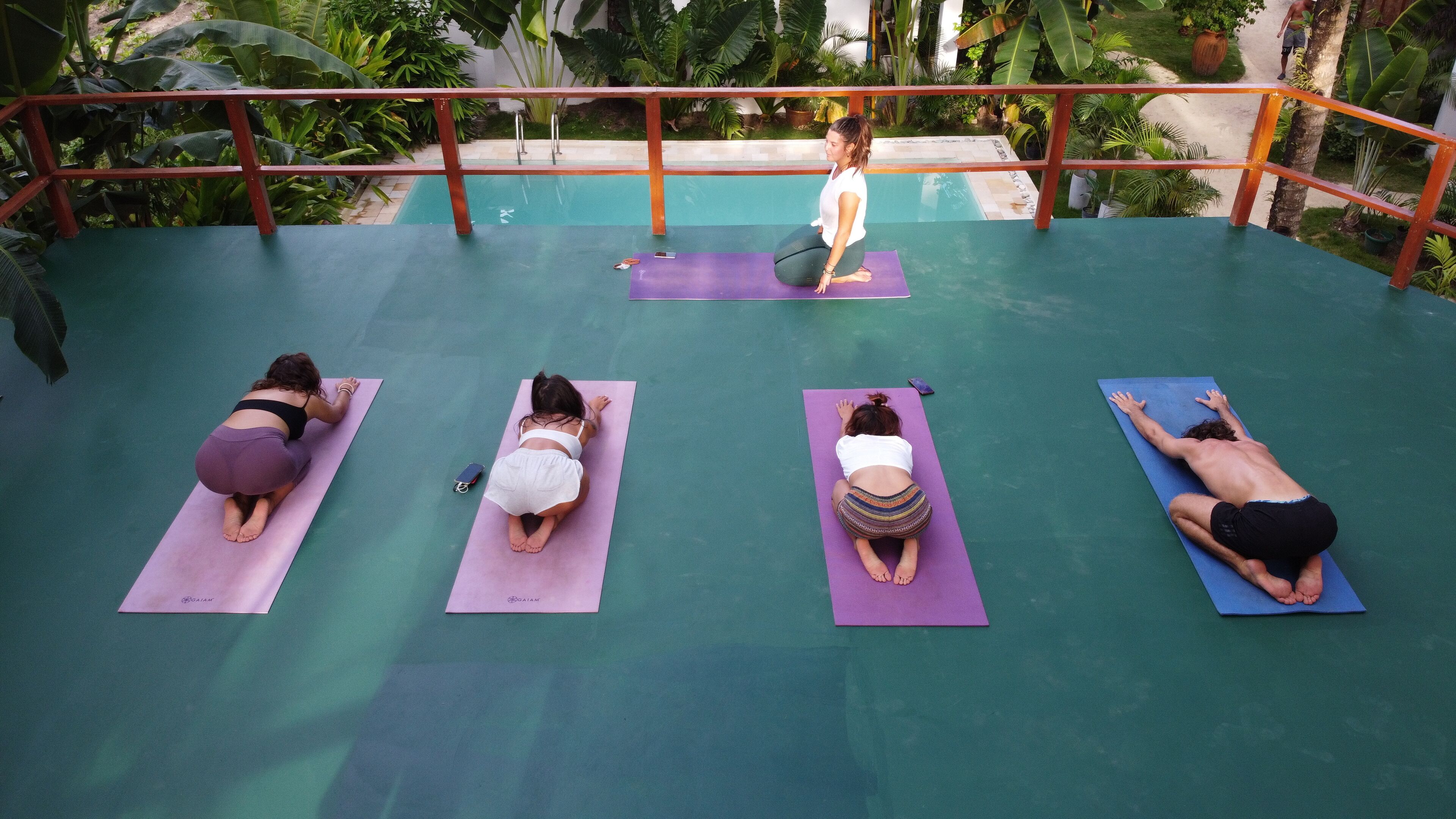 A group of people on yoga mats in an outdoor setting with a pool and lush greenery in the background.