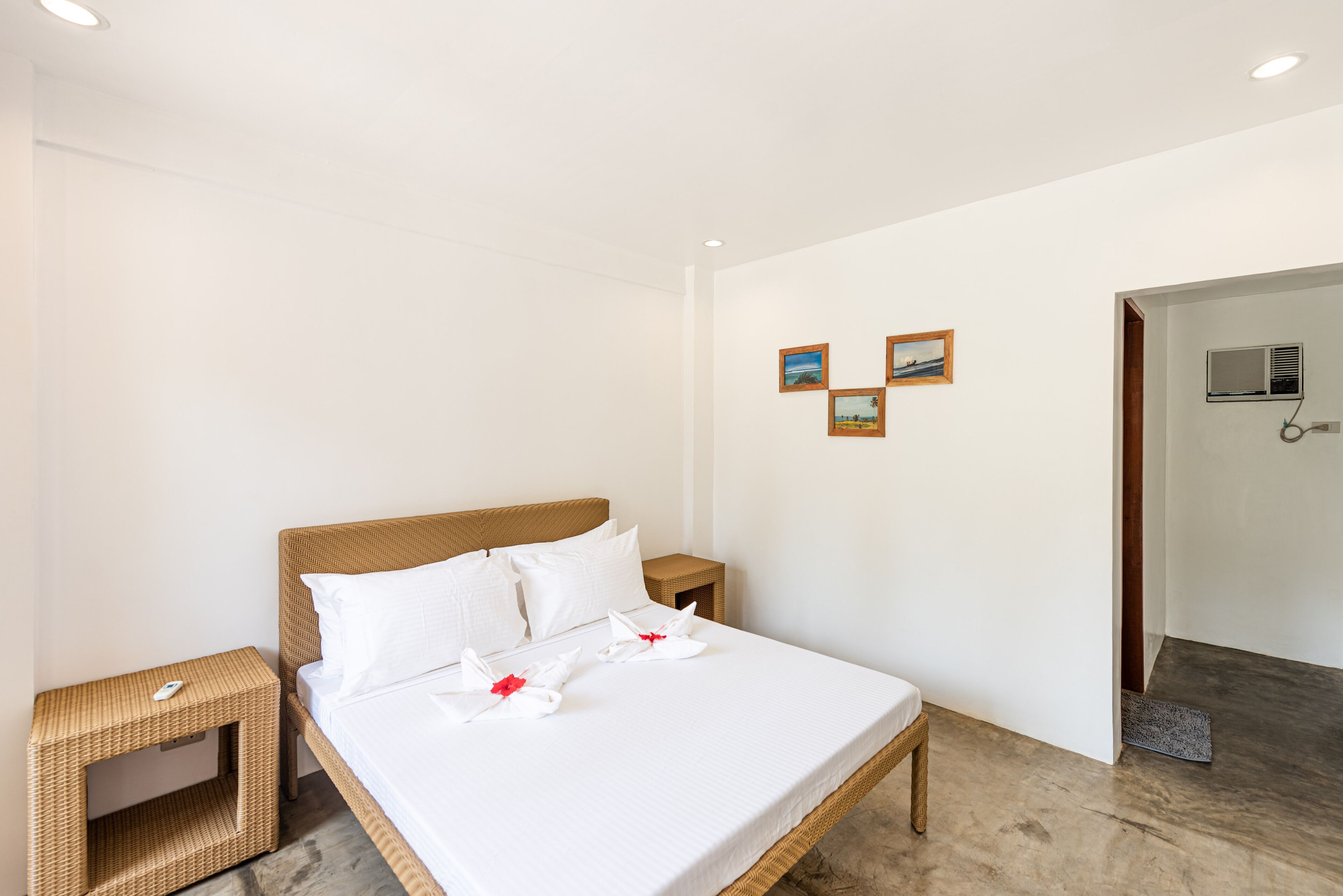 Een net opgemaakt bed met witte lakens en kussens, een klein rieten nachtkastje met een afstandsbediening, en een ingelijst schilderij aan de muur.