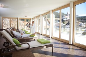 Zona de spa con sillones reclinables, un masajista y grandes ventanales con vistas a un pueblo e iglesia.