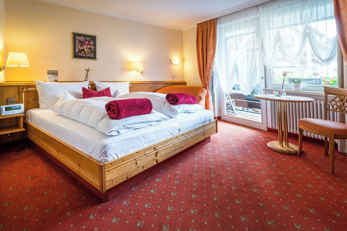 Habitació d'hotel amb llit de fusta, catifes vermelles, dues làmpades al costat del llit, una taula petita i una finestra amb cortines.