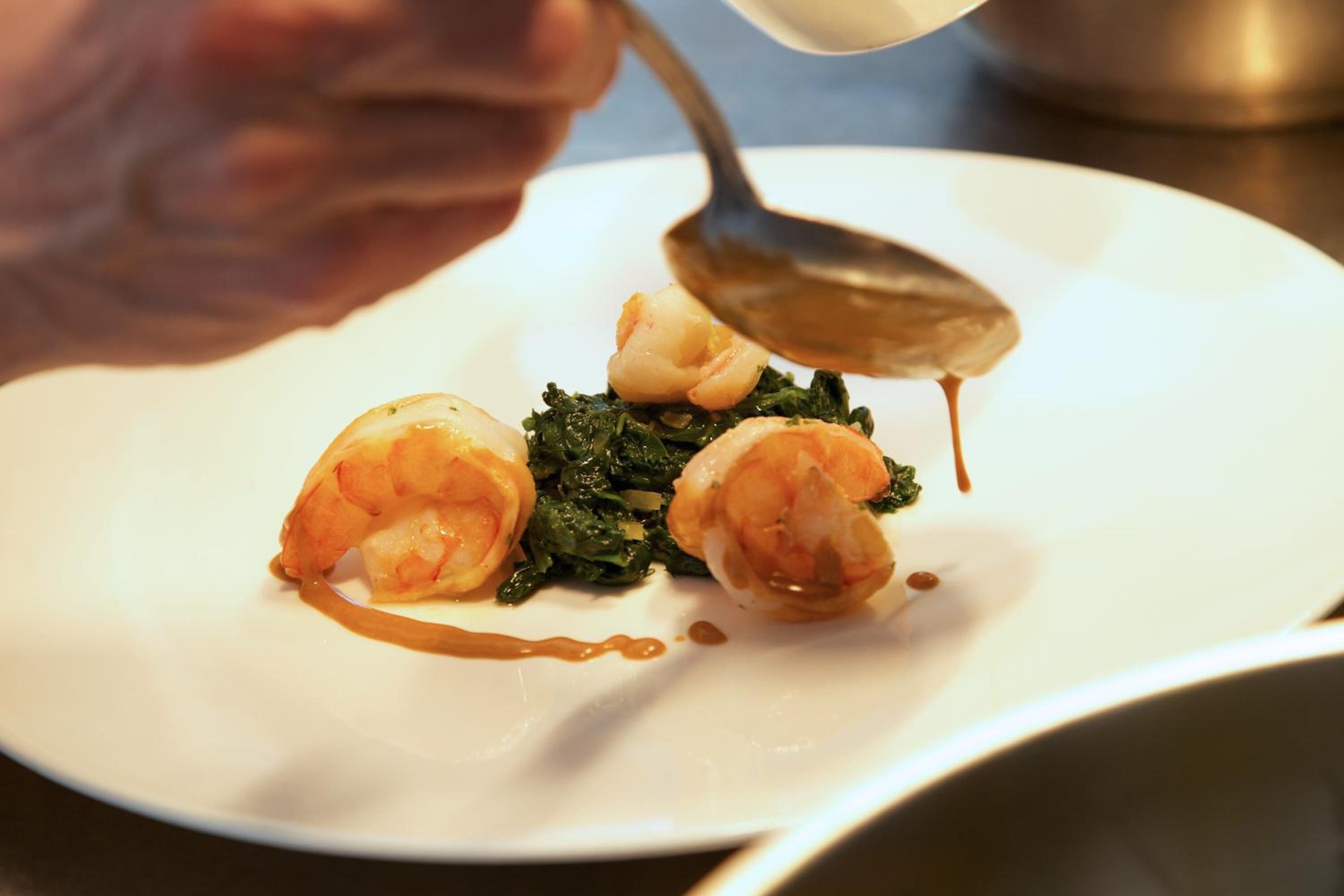 A person pouring sauce onto a plate with shrimp and greens.