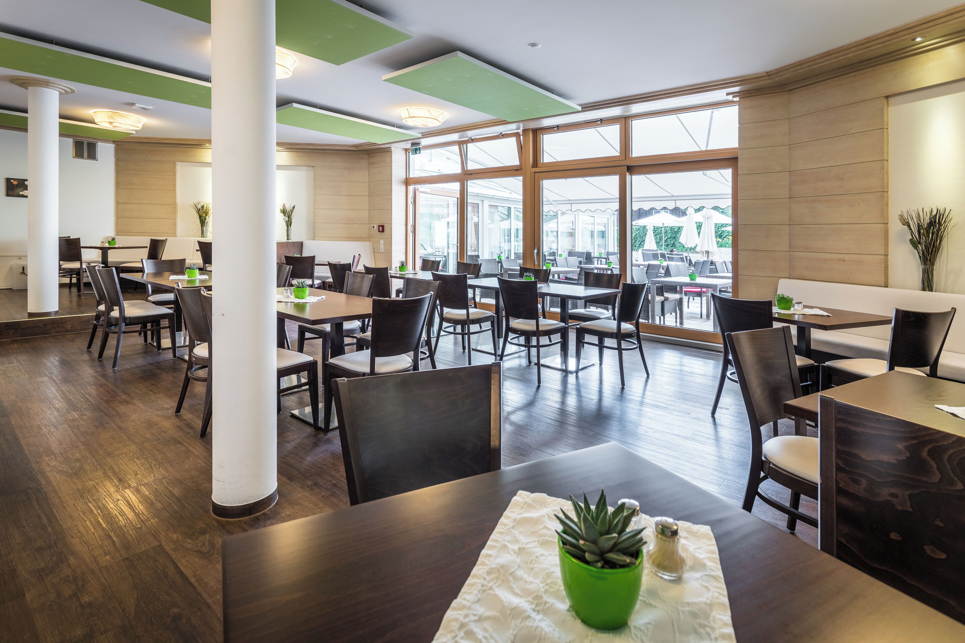 Un restaurante moderno con suelo de madera, paneles de techo verdes y grandes ventanales que ofrecen vistas al exterior.