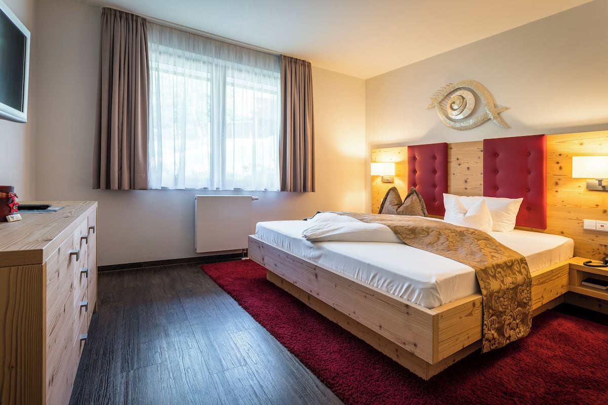 Habitació d'hotel amb llit de fusta, una catifes vermella, un taulell de vestir de fusta, finestra amb cortines i un objecte decoratiu penjat a la paret.