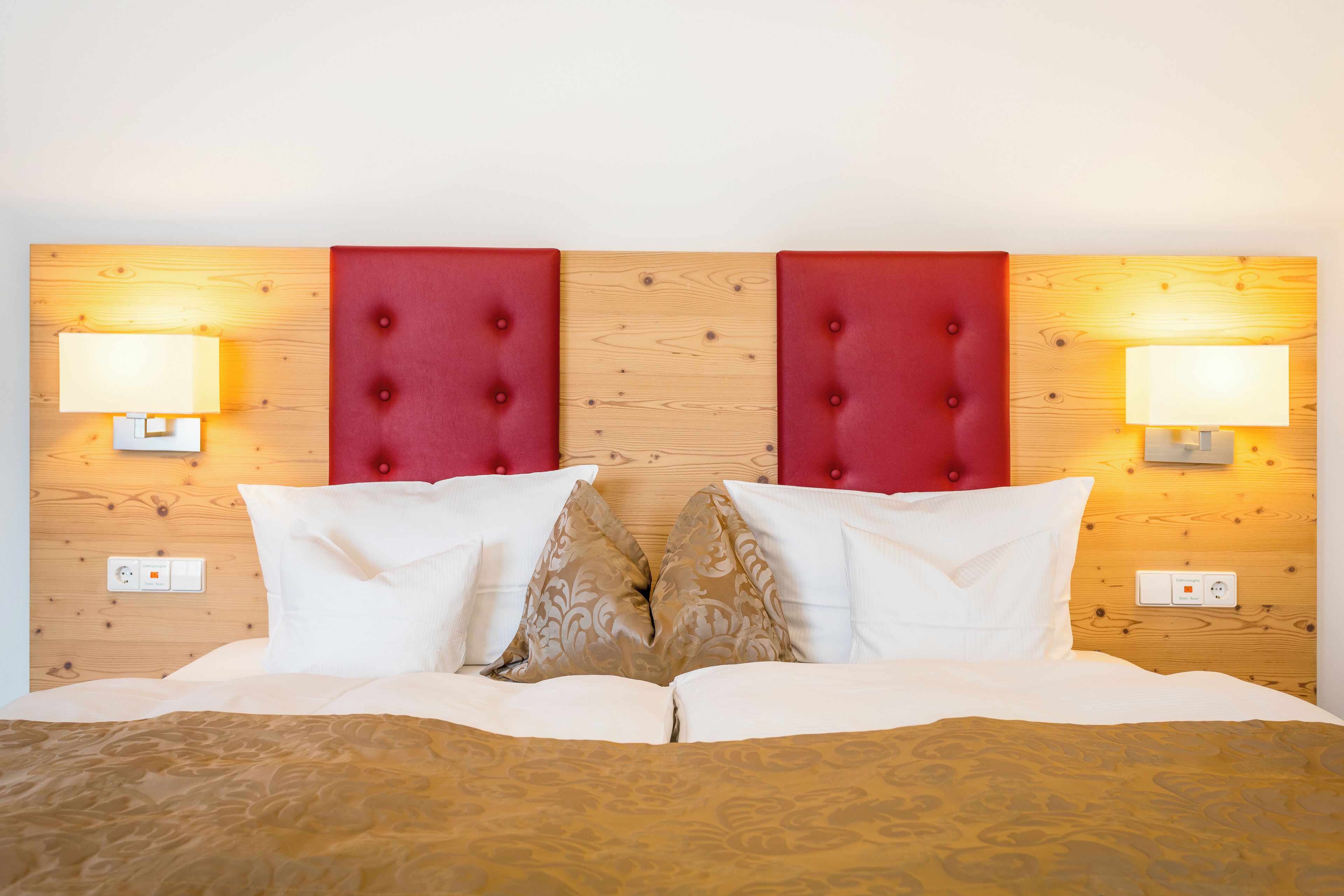 Una camera d'albergo con una testiera in legno, due lampade ai comodini, un letto con cuscini bianchi e decorati e una presa elettrica sulla parete.