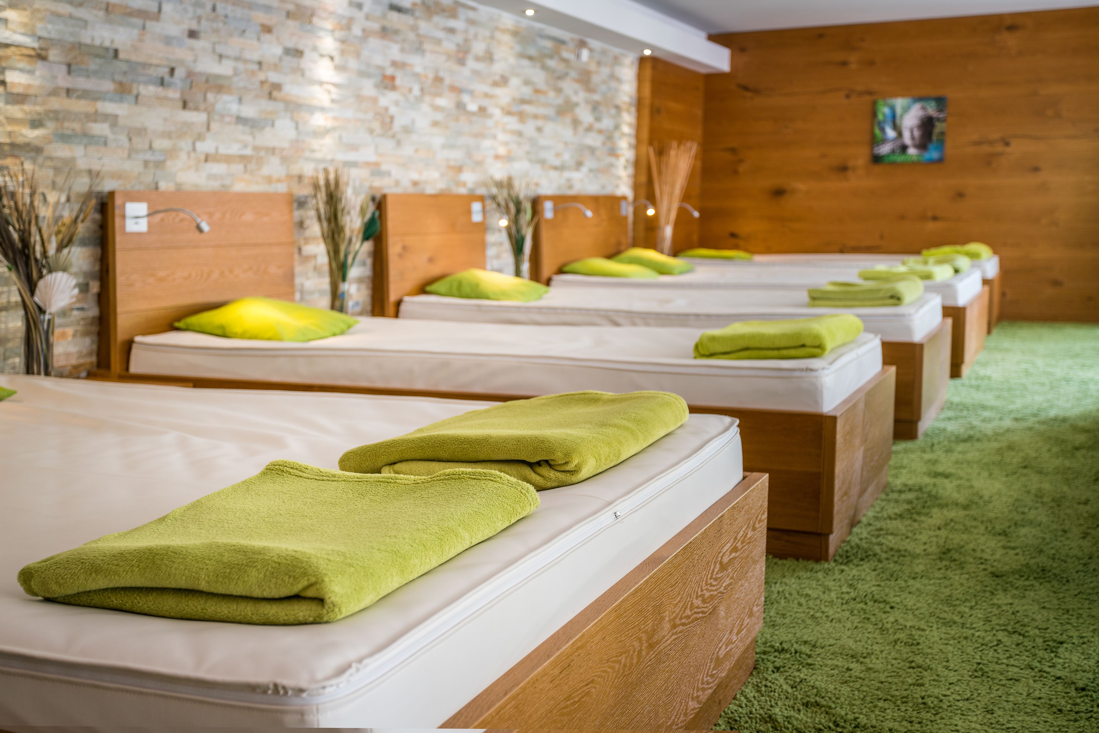 A row of neatly made beds with green towels, wooden headboards, and a brick wall in the background.