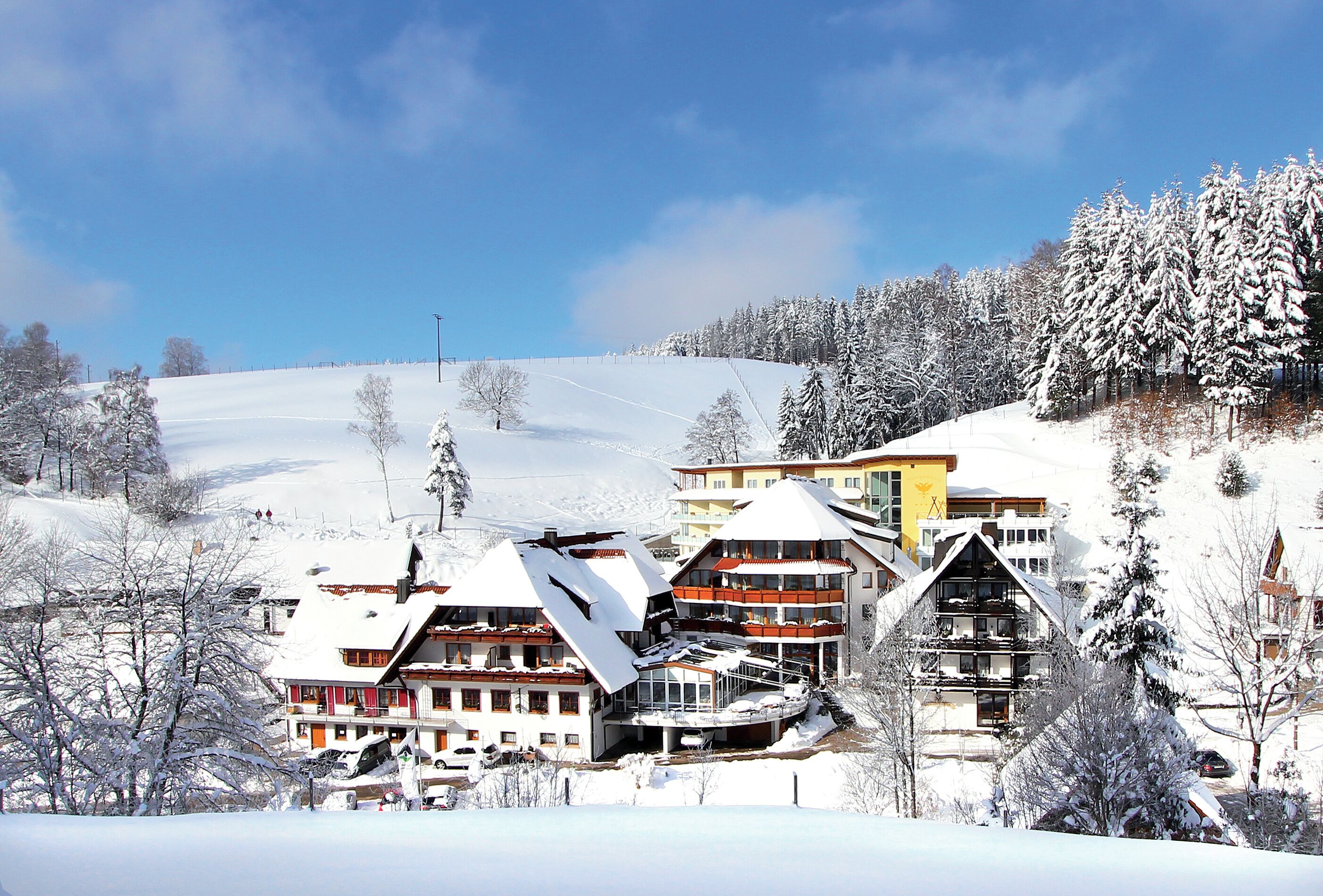 A snow-covered mountain resort with multiple buildings, including a hotel with balconies and a ski lift in the background.
