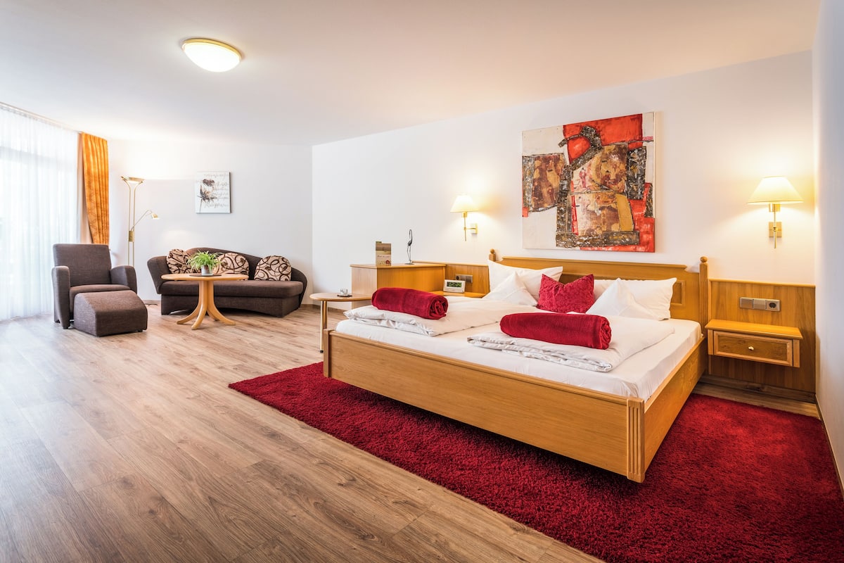 Habitació d'hotel moderna amb llit de fusta, catifes vermelles i dues taules de nit amb làmpades.