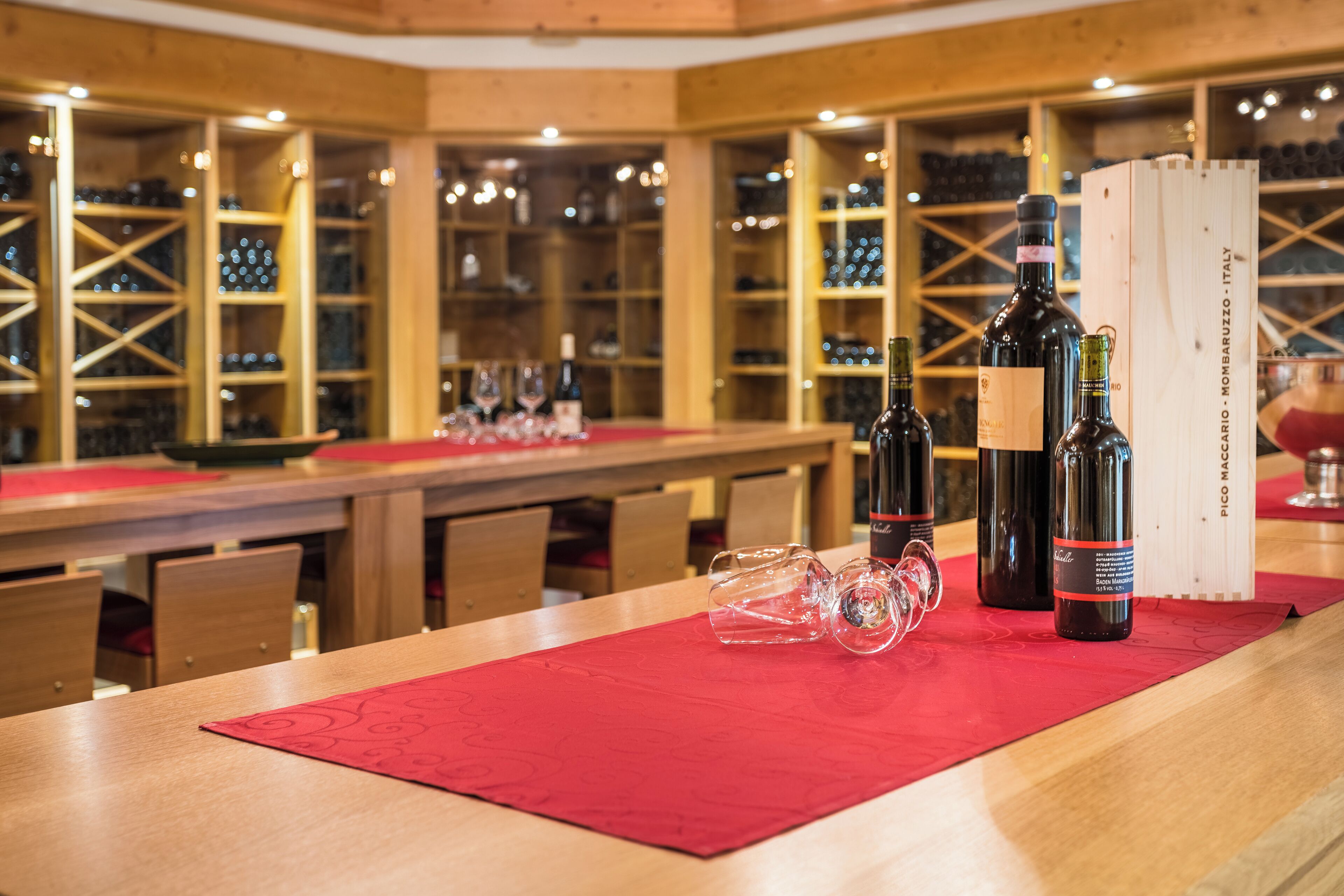 A wine tasting room with wooden shelves, a long table with a red placemat, wine bottles, and glasses.
