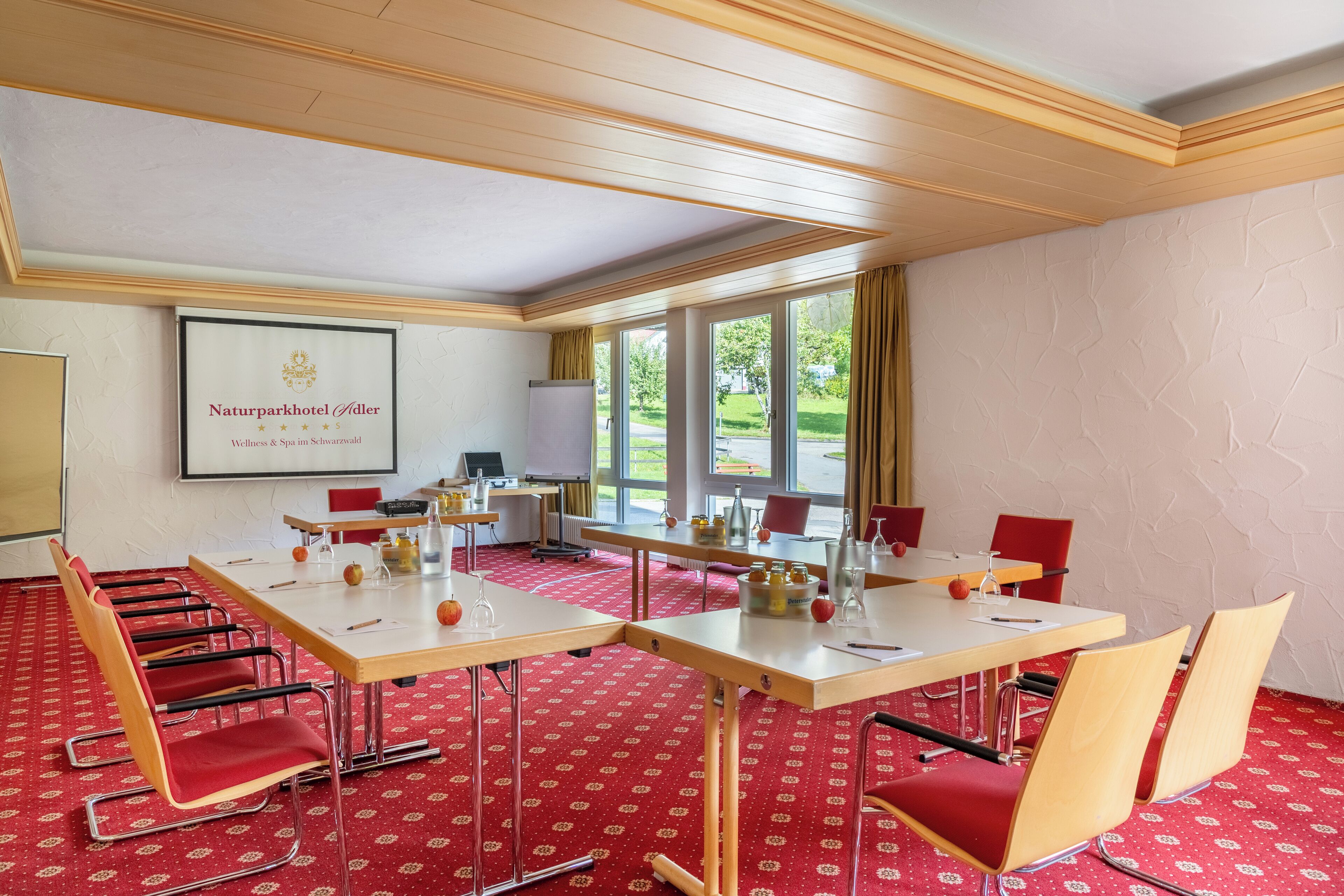 Een congreszaal met een grote tafel, rode stoelen en een projectiescherm waarop het logo van Naturparkhotel Adler staat.
