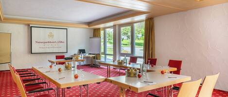 Een congreszaal met een grote tafel, rode stoelen en een projectiescherm waarop het logo van Naturparkhotel Adler staat.